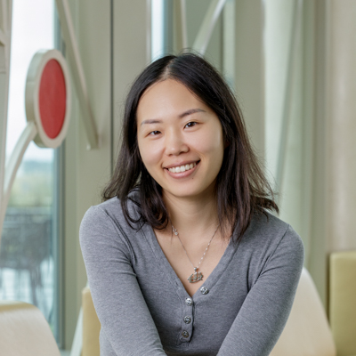 An Asian woman smiling, with short black hair, wearing a grey blouse and beige pants.