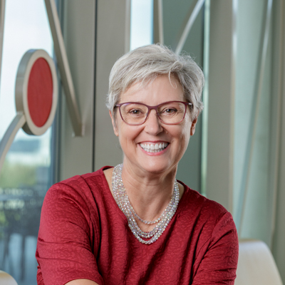 Portrait of Sue Bruns, a white woman smiling with short gray hair, glasses, and a red dress.