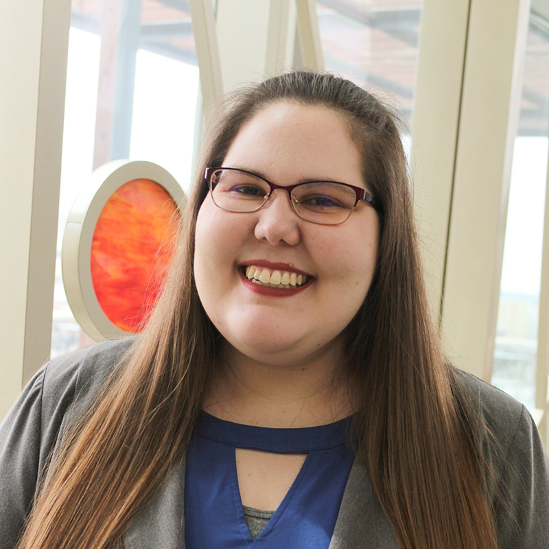 A white woman in a blue top smiling and standing in front of the 100 Women Wall of Honor