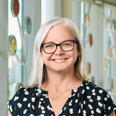 A white woman smiling, with blonde, straight hair, wearing black-frame glasses and a black shirt with polka dots.