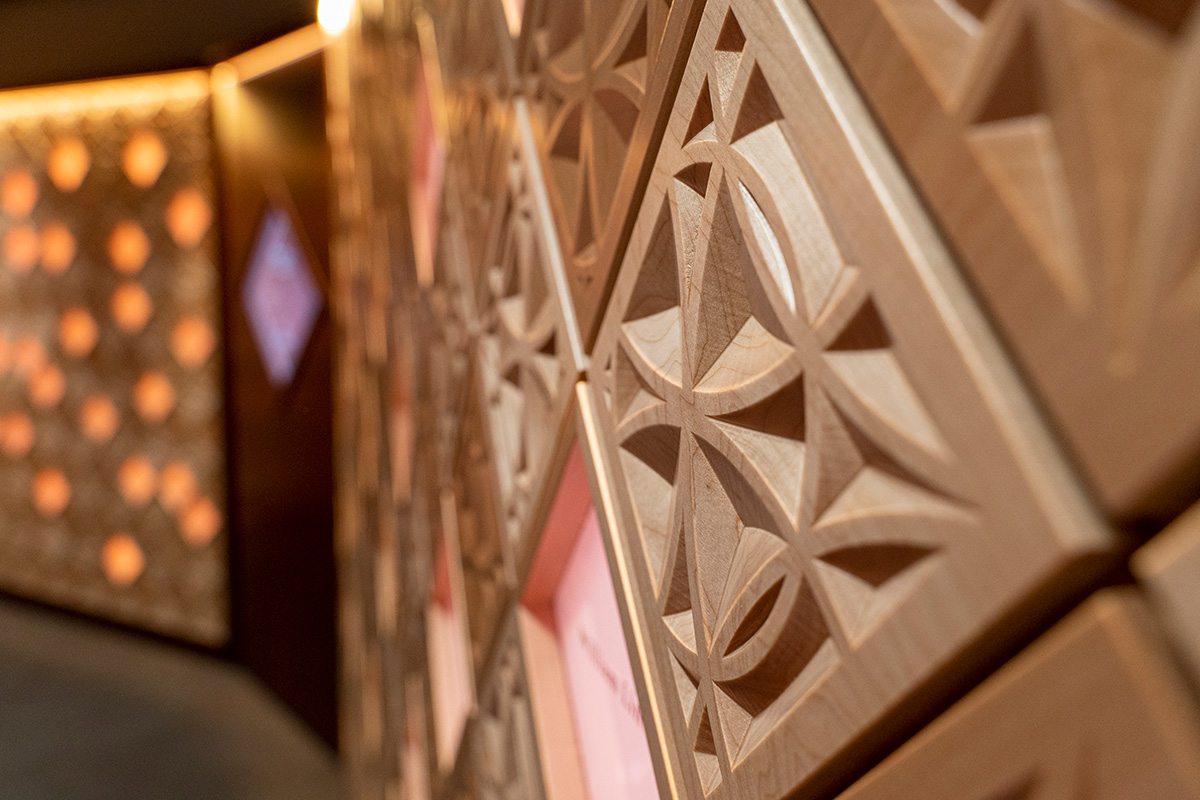 Close up of the wall in Ember Hall, composed of ornately carved, wooden square diamond shapes. Amongst the tiles are glowing diamonds lit in colors of pink and organge.