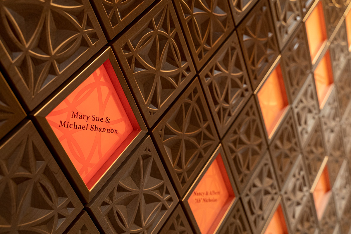Close up of the wall in Ember Hall, composed of ornately carved, wooden square diamond shapes. Amongst the tiles are red, glowing diamonds displaying donor names