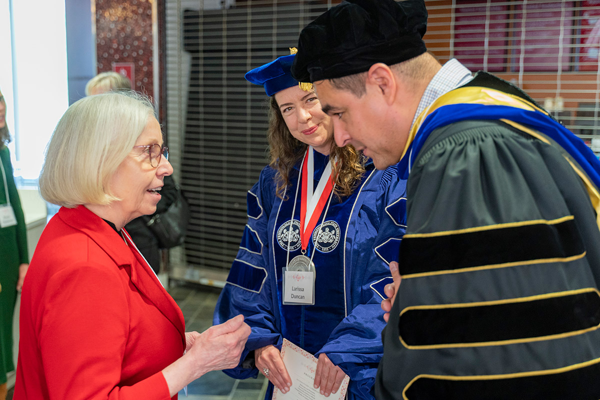 Three people face each other in conversation. Two are faculty members dressed in academic regalia