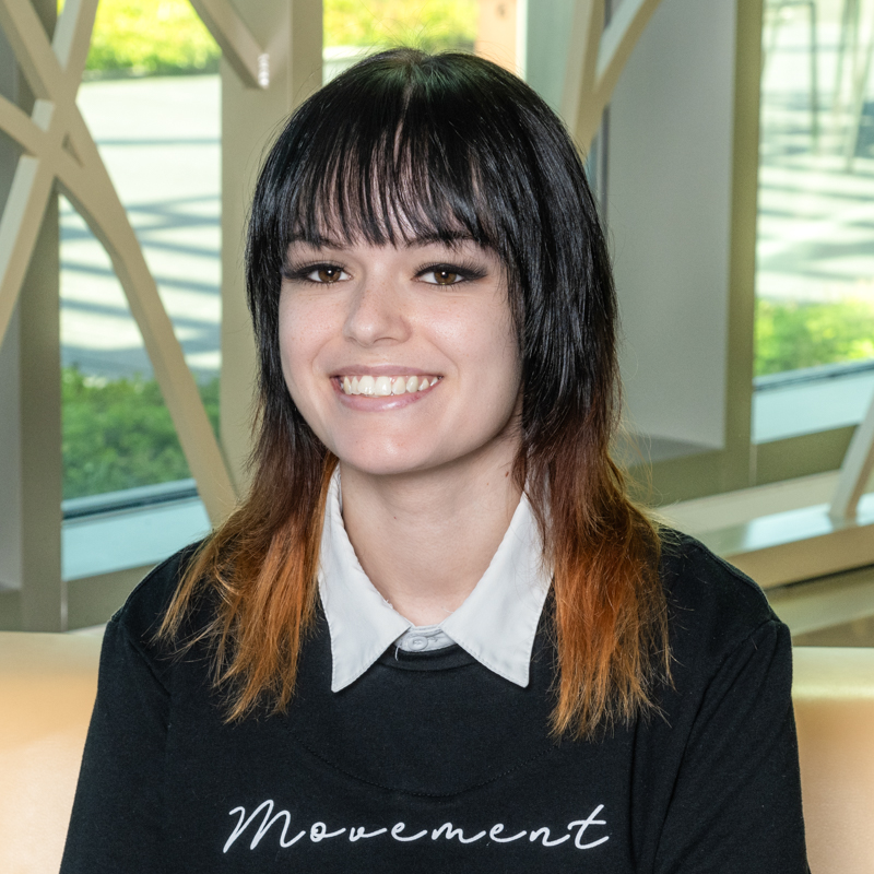 A white woman smiling, with black ombre to brown straight hair wearing a black shirt with a white collar.