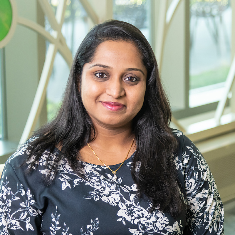 Professional portrait of Chandni Anandha Krishnan in front of a decorative window with bright colored glass circles and curved lines.