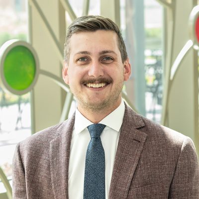 A white man smiling, with short brown hair and a mustache, wearing a navy blue tie with a suit jacket.