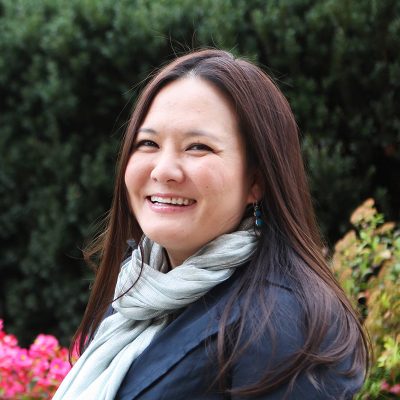 An Asian woman smiling, with shoulder-length brown hair, wearing a blue neck scarf and jacket.