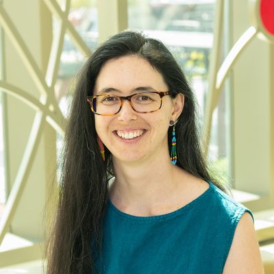 A woman smiling, with long, straight, dark hair, wearing rectangle glasses, a sleeveless teal top, and long earrings.