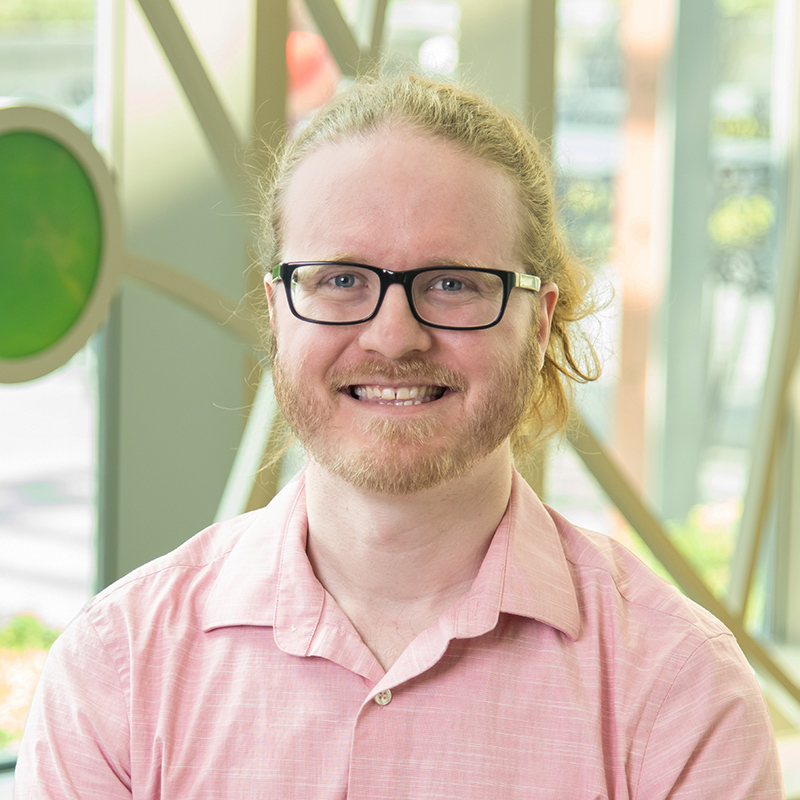 A man smiling, with blond hair and facial hair, wearing black frame glasses and a pink collared, button-up shirt.