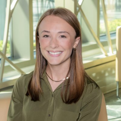 A white woman smiling, with mid-length auburn hair, and wearing an olive green button up shirt and gold jewelry.
