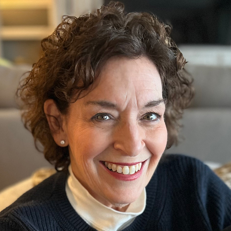 A woman with dark brown hair, dressed in a blue sweater, who is happy and welcoming.