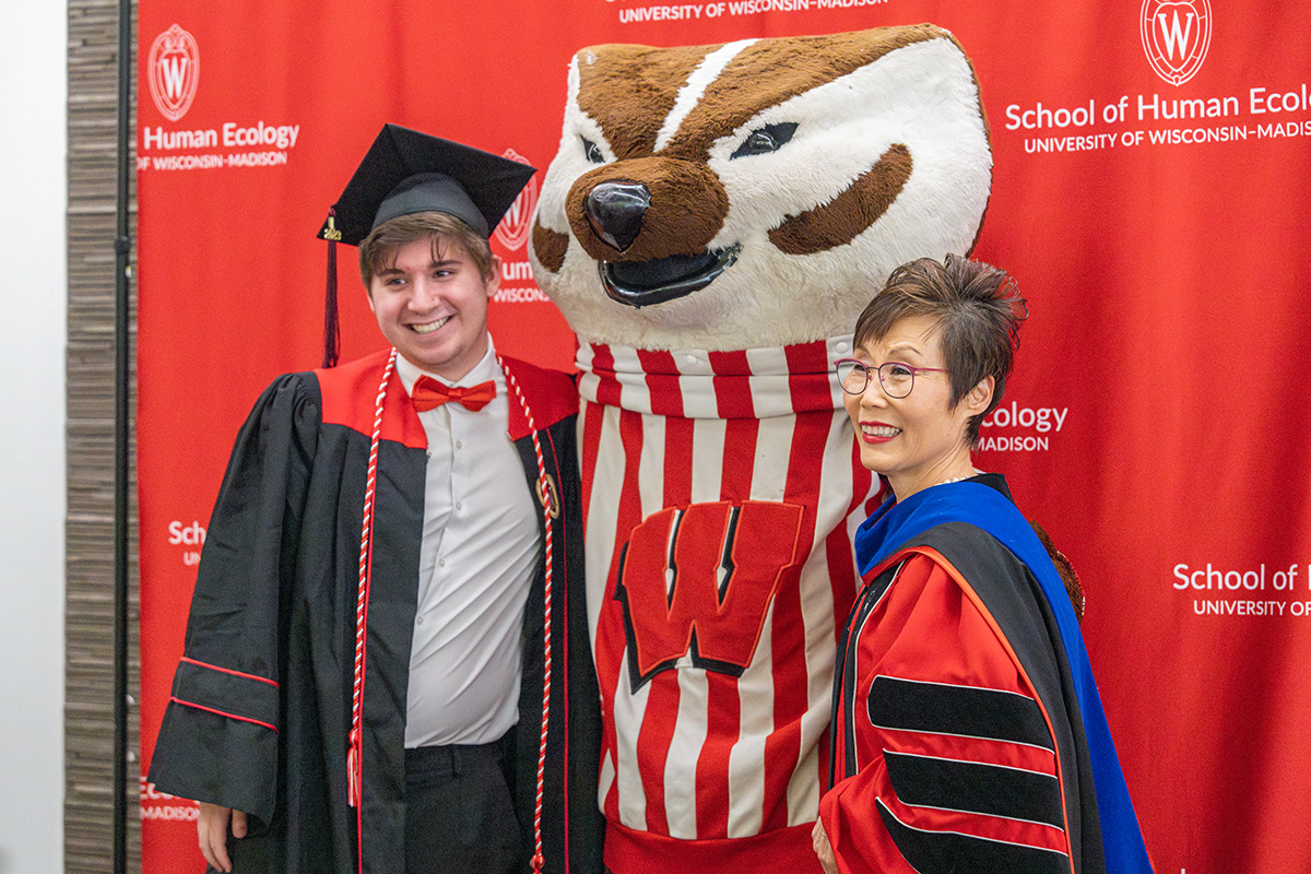 Undergraduate student and dean in graduation attire pose with Bucky Badger in front of a red backdrop.