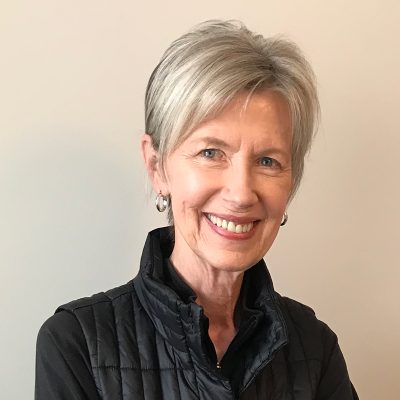 white woman, smiling, with blonde-gray hair, wearing black vest and earrings, on a gray background