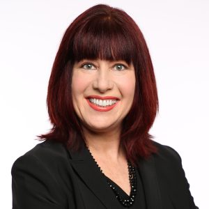 A woman smiling with a friendly expression, with reddish-brown hair and wearing a black suit