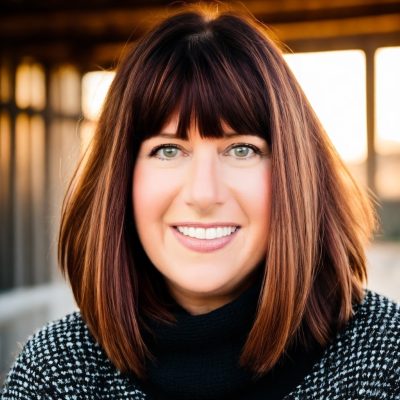 A woman with shoulder-length reddish-brown hair and bangs smiles at the camera. She is wearing a black turtleneck and a black-and-white patterned sweater. The background is softly blurred, suggesting an outdoor setting with warm sunlight.