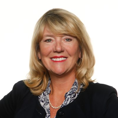 A white woman smiling, arms at her sides, with blonde shoulder length hair and wearing a navy blue suit.