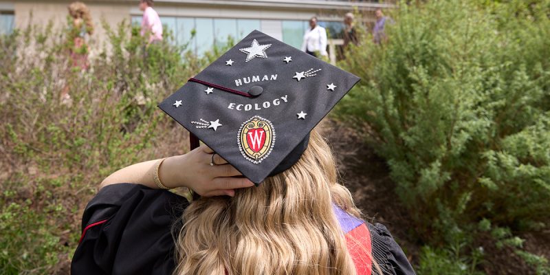 Graduate with blonde hair wears a graduation cap that is decorated in stickers that spell out "Human Ecology"