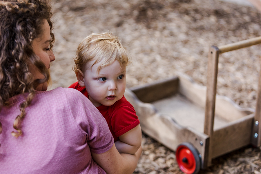 An adult holds a small child. The child is looking over their shoulder and the adult is looking at them. There is a toy wagon in the background.