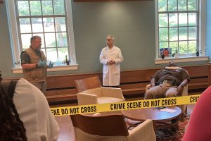 Three men, two standing and one seated, are pictured behind yellow crime scene tape in the foreground.