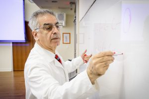 A man with short, gray hair wearing glasses, a white lab coat and a red tie writes on a whiteboard with a red dry erase marker.