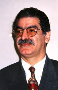 A man with short, dark hair wearing glasses, a white shirt, dark jacket and patterned red tie looks off camera.
