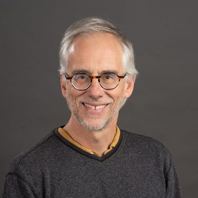 A white man smiling, with gray hair, wearing glasses and a dark gray sweater.