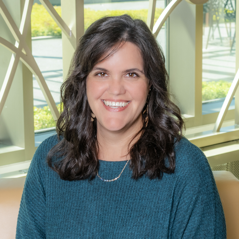 A white woman with brown hair and eyes wearing a teal sweater and gold earrings smiles in front of a large window.