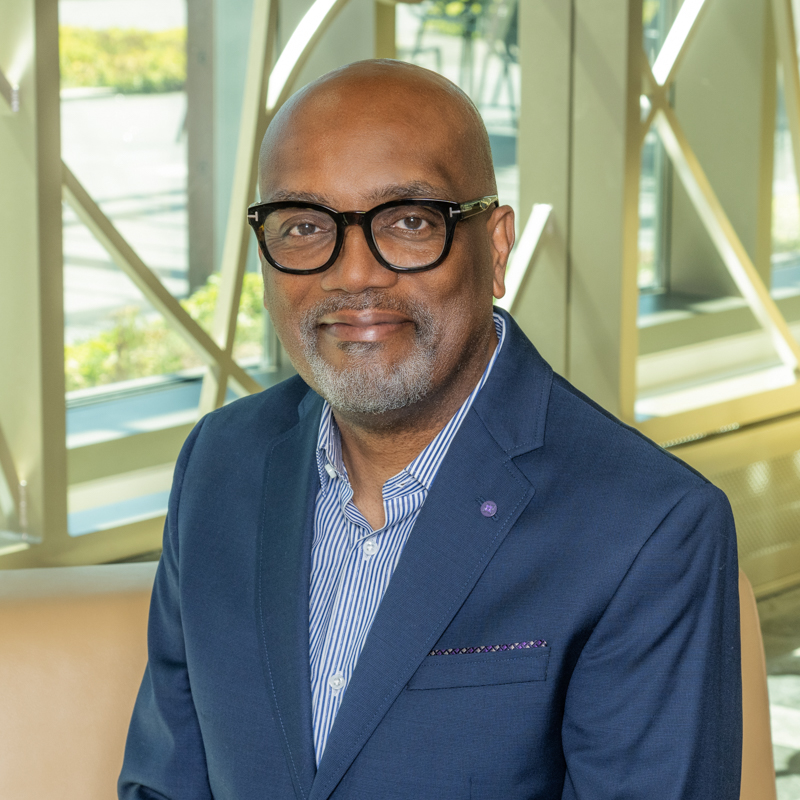A Black bald-headed man with eyeglasses wearing a blue and white striped shirt and a blue jacket.