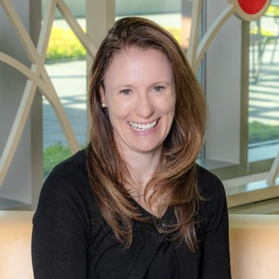 A white woman smiling, with brown straight hair, wearing a black sweater.
