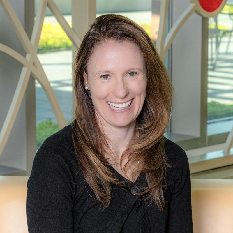 A white woman smiling, with brown straight hair, wearing a black sweater.