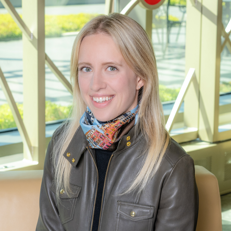 A white woman smiling, with blonde, long hair, wearing an olive jacket.
