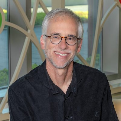 A white man smiling, with gray hair and a close-cropped beard, wearing glasses and a black shirt.