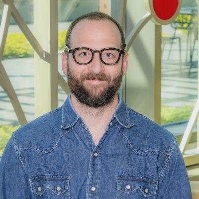 A white male smiling with short hair, glasses, and a beard, wearing a blue denim shirt.