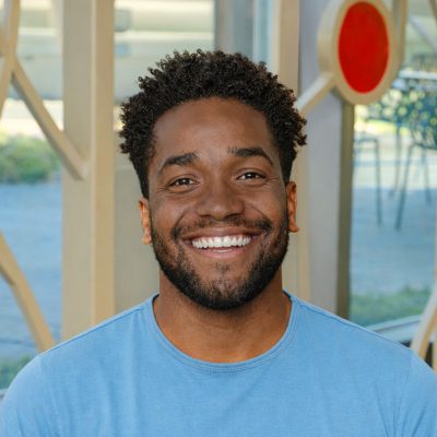 A black man smiling, with black, curly hair, wearing a light blue shirt.