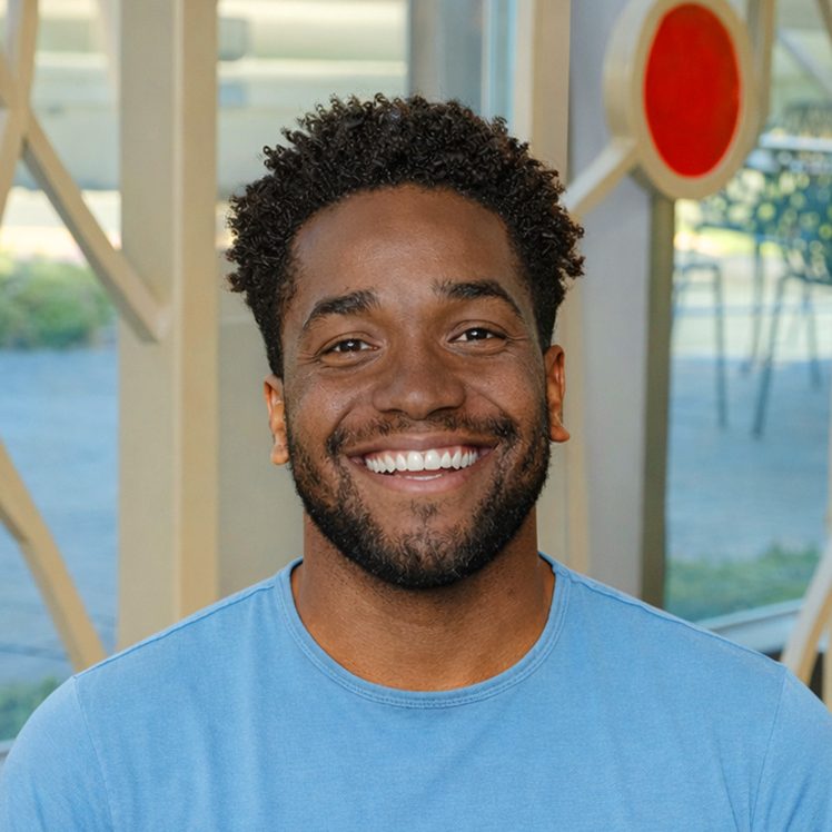 A black man smiling, with black, curly hair, wearing a light blue shirt.