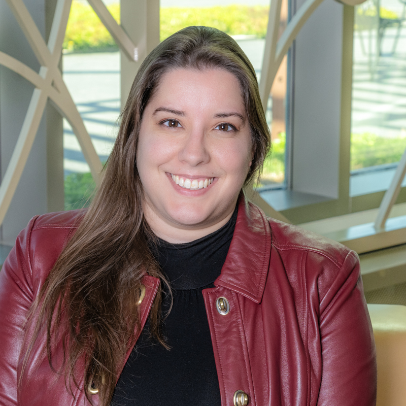 A white woman smiling, with long brown hair, wearing a black shirt and red coat.