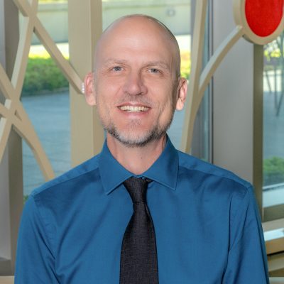 A white man smiling, wearing a blue shirt with a black tie.