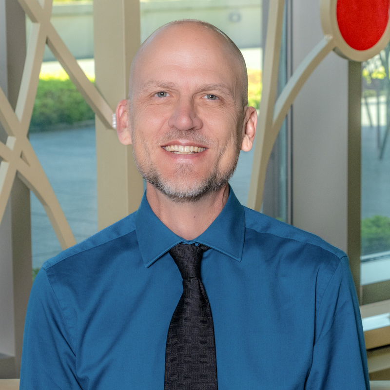 A white man smiling, wearing a blue shirt with a black tie.