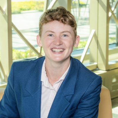 A white person with a big smile and short, dark blonde hair, wearing a light shirt and a navy blazer.