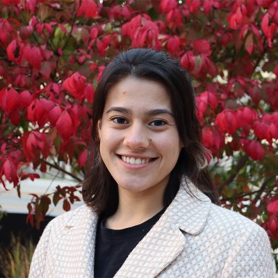 A young woman smiling, with straight, shoulder-length, dark brown hair, wearing a beige blazer against a backdrop of red foliage.
