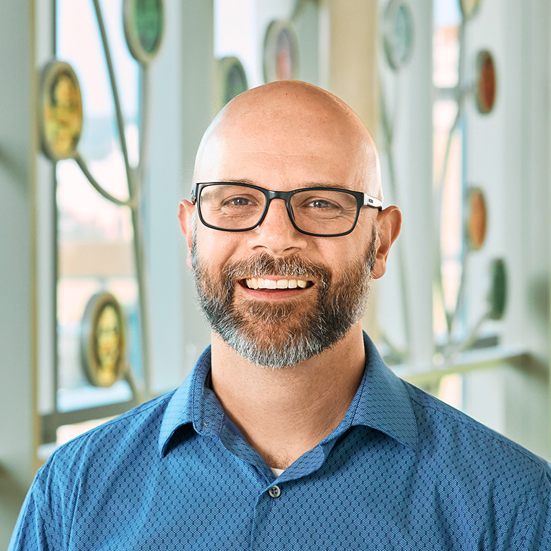 A white bald man with black glasses and a beard, wearing a blue shirt and smiling.