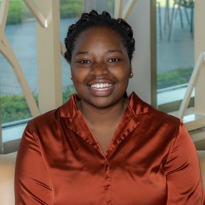 A black woman smiling, with maroon and black braids, wearing a copper satin shirt and silver earrings.