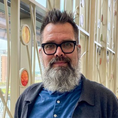 A white man with a beard and glasses, smiling, wearing a blue shirt and a grey sweater.