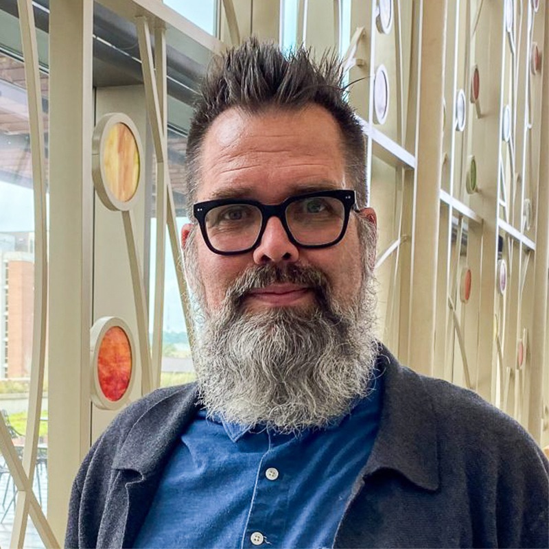 A white man with a beard and glasses, smiling, wearing a blue shirt and a grey sweater.