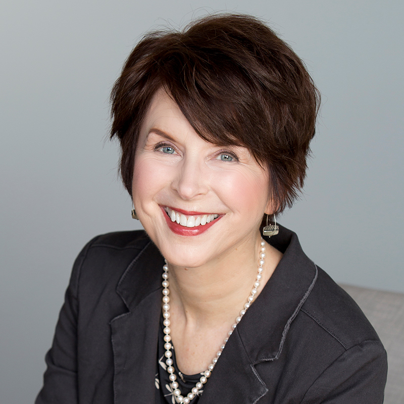A white woman smiling, with short auburn hair, wearing a blue blazer and a pearl necklace with silver charm.