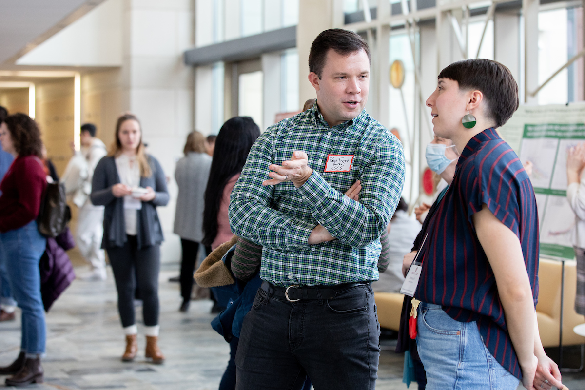 Two people, turned toward one another, engage in a conversation while others mill about in the background.