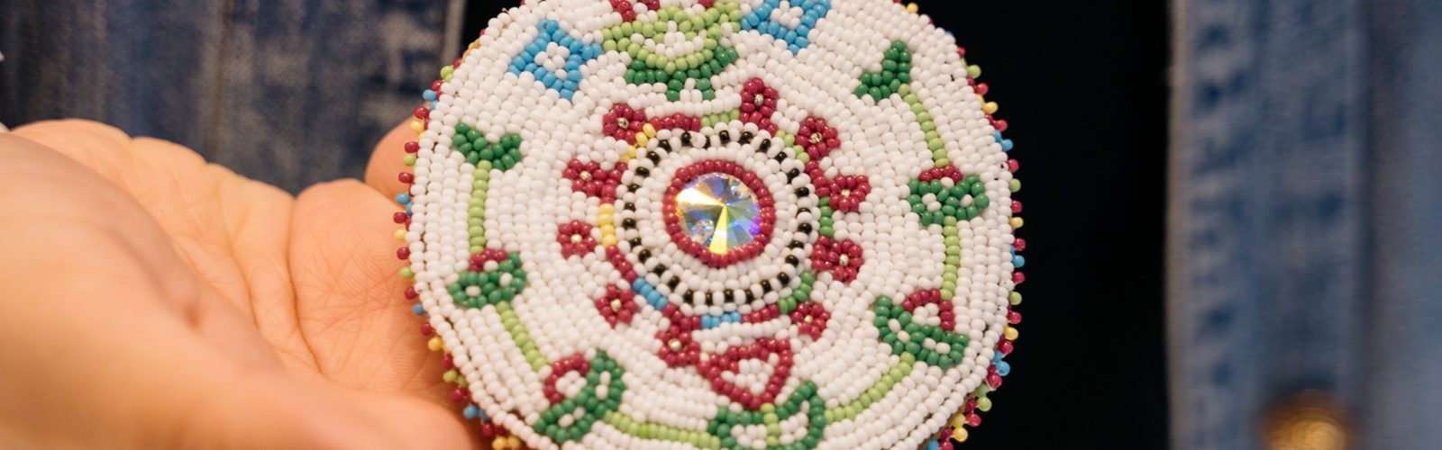 Close-up of a hand holding a circular beaded medallion, which is mostly white with a colorful growing flower motif.