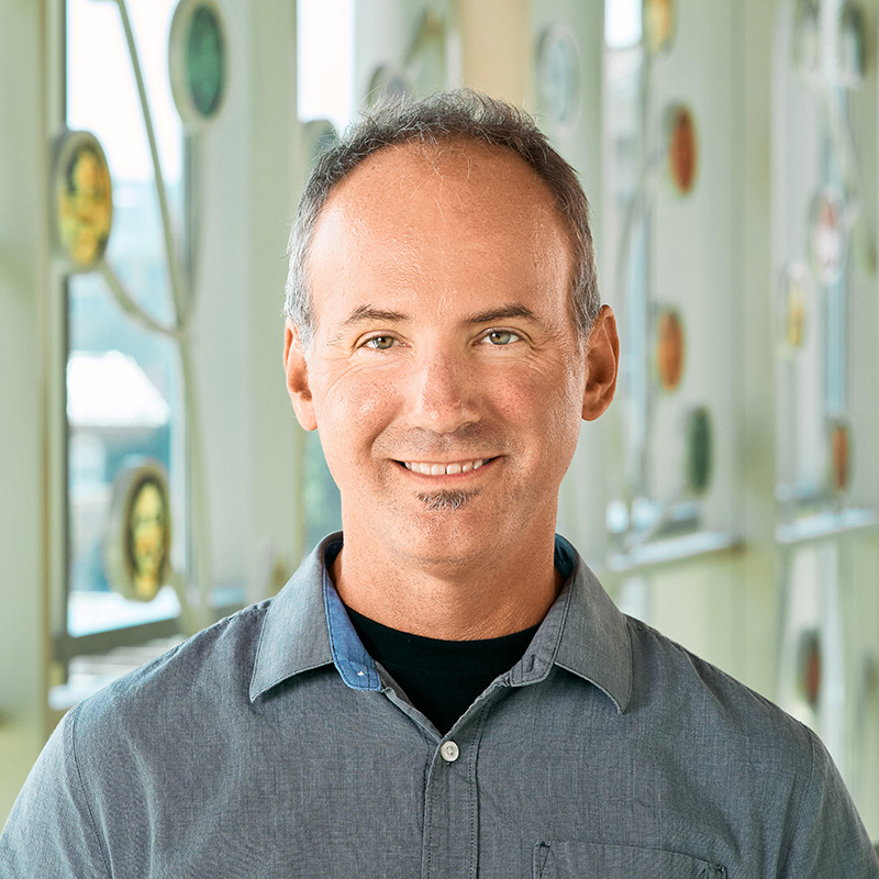 A white man smiling, with short, grey hair, wearing a grey button-up shirt.