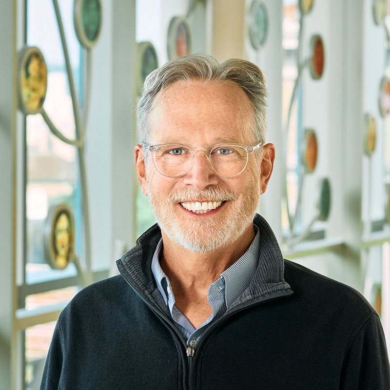 A white man with gray hair, glasses, and trimmed beard, smiling wearing a light blue shirt and a dark sweater.