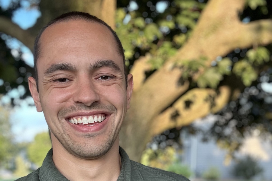 A male with short dark hair in a green shirt smiling at the camera with a tree in the background.